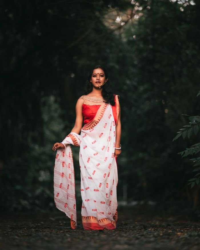 about-us-02 A young woman poses elegantly in a traditional sari amidst a serene forest path.