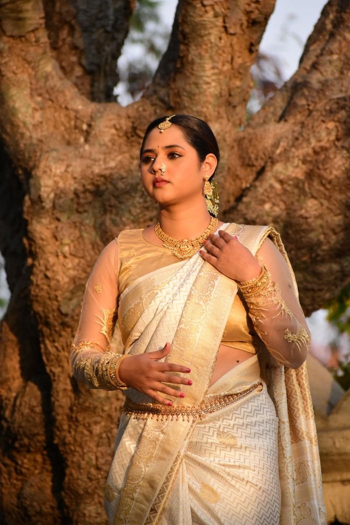 collection-04 A woman in a golden saree stands gracefully by a tree, exuding cultural elegance.